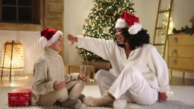 Sevgi dolu gülümseyen anne oğluna Noel hediyesi sunuyor, modern rahat ışık odasında yerde oturuyor ve Noel ağacında çelenk takıyor. Yüksek kalite 4k görüntü