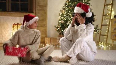 Aydınlanmış Noel ağacı olan rahat bir oturma odası. Orta yaşlı, 9-10 yaşlarında, Noel Baba şapkaları ve sıcak ev kıyafetleriyle yerde oturan bir anne ve oğlu. Yüksek kalite 4k görüntü