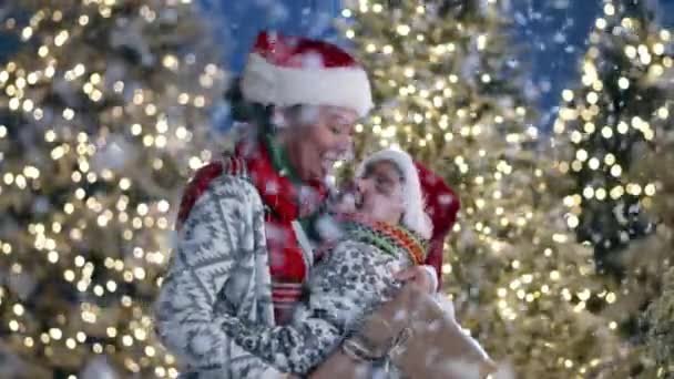 ハッピーマザー サンタの帽子の息子 前景にふわふわの雪片で抱き合う装飾のセーター 背景に照らされたクリスマスツリー 高品質の4K映像 — ストック動画