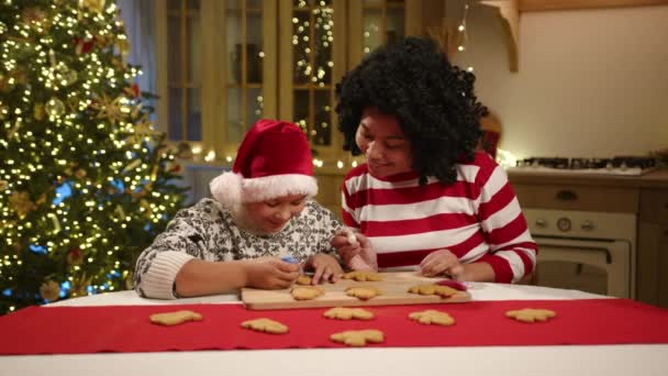 可爱的母亲和小儿子坐在厨房桌子旁 画着自家做的姜饼 现代厨房装饰着闪亮的花环和圣诞树 高质量的4K镜头 — 图库视频影像