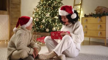 Noel Baba şapkalı, beyaz pijamalı güzel siyah kadın hediye paketini açıp oğlunun yanağına dokunduktan sonra hoş bir sürprizle karşılaşır. Noel süsleri. Yüksek kalite 4k görüntü