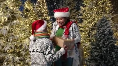 9-10 yaşlarında Noel Baba şapkalı bir çocuk annesine Noel kutusunu veriyor yoğun kar yağışı ve parlak çelenklerle süslenmiş kozalaklı büyülü bir yerde sarılıyor. Yüksek kalite 4k görüntü