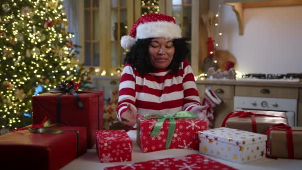 笑容满面的黑人女人戴着圣诞礼帽坐在餐桌旁 桌上摆满了圣诞礼物 厨房装饰华丽 圣诞树灯火通明 盒子开封 高质量的4K镜头 — 图库视频影像