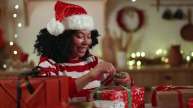 Gülümseyen kıvırcık saçlı Afro-Amerikan kadın Noel şapkası içinde hediyeleri sevgi ve özenle paketliyor, parlak çelenklerle süslenmiş rahat bir odada oturuyor. Yüksek kalite 4k görüntü