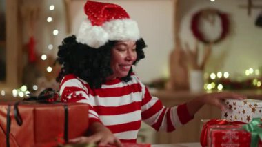 Noel şapkalı orta yaşlı bir Afrikalı kadın festival süslemeli bir odada oturuyor, hediyelerle dolu kutuyu seçiyor, açtıktan sonra mutluluk çığlıkları atıyor. Yüksek kalite 4k görüntü