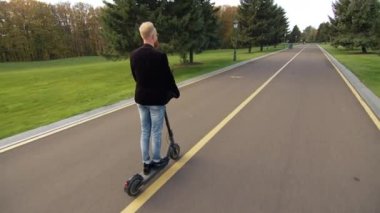 Uzun bisiklet yolu, yeşil çimenli uçsuz bucaksız çimenler, yeşil ağaçlar, sokak lambaları ve ziyaretçiler için banklar. Parkta başka bir scooter sürücüsü. Yüksek kalite 4k görüntü