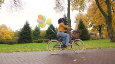 Retro bisikletli renkli bir kadın sepetinde balonlar ve çiçeklerle parkta dolaşırken mutluluk ve özgürlükten kahroluyor. Sonbaharda doğanın güzelliği. Yüksek kalite 4k görüntü