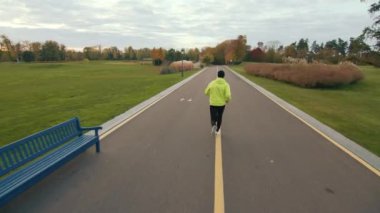 Güneşli bir sonbahar gününde yeşil çimenler, çok renkli ağaçlar ve çalılarla ıssız güzel bir parkta spor kıyafetleriyle koşan uzun boylu bir erkek koşucunun arka görüntüsü. Yüksek kalite 4k görüntü