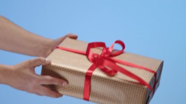 Erkek eller, ince kırmızı kurdeleli büyük bir hediye paketi uzatıyor ve mavi arka planda bakımlı kadın ellerine eğilip gözden kayboluyor. Yüksek kalite 4k görüntü