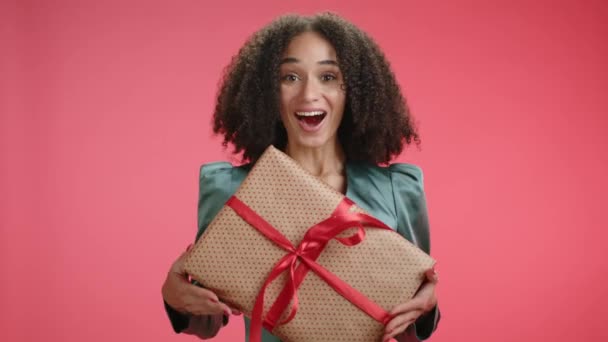 喜ばしいカーリー髪のブルネットは 赤い背景に隔離された認識できない人からギフト包まれた箱を得ます 非常に幸せな女性は贈り物で箱を揺ります 高品質の4K映像 — ストック動画