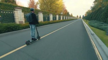 Elctro scooter kullanan uzun boylu bir adam orta hızda düz çizgi üzerinde. Yeşil çimenlikli güzel bir peyzaj, yemyeşil ağaçlar, yol boyunca çalılar. Yüksek kalite 4k görüntü