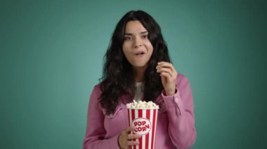 Güzel kız yüzünde önyargılı bir gülümsemeyle bir film izliyor. Elinde patlamış mısır tutuyor ve yiyor. Pembe elbiseli kız mavi arka planda. Yüksek kalite 4k görüntü