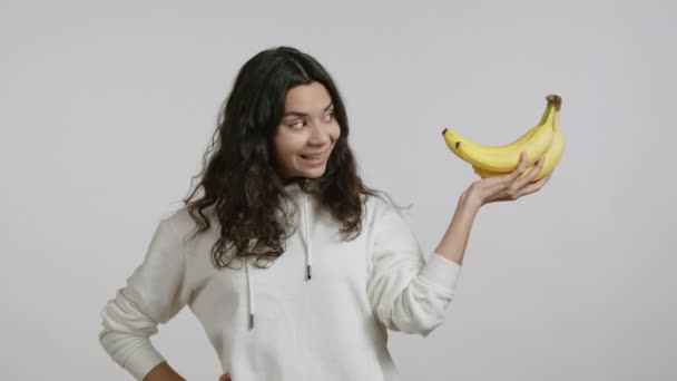 美しい女の子が黄色いバナナを展示しています 彼女は白いバックグラウンドに白いパーカーをしています 少女は幸せで喜んでいる 高品質の4K映像 — ストック動画