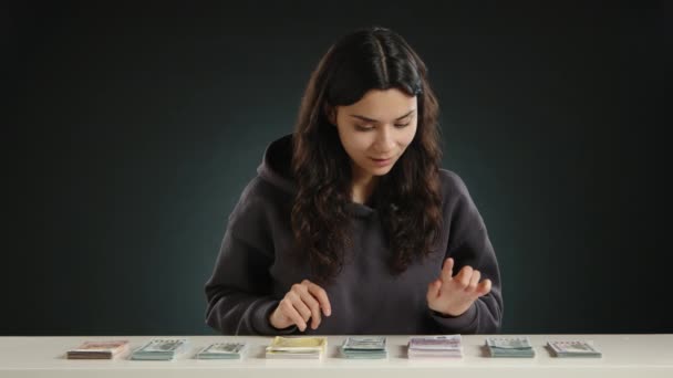 暗いバックグラウンドのダークフーディーの若い女の子がテーブルの上のドル紙幣の積み重ねを調べています 高品質の4K映像 — ストック動画
