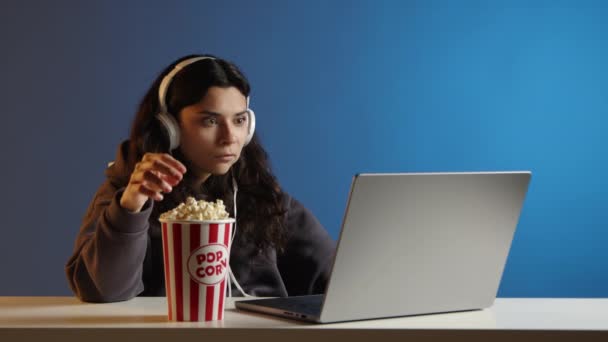 一个戴耳机的漂亮女孩在笔记本电脑上看电影 吃爆米花 她在等待着什么 但后来她对自己在笔记本电脑屏幕上看到的东西感到失望 — 图库视频影像