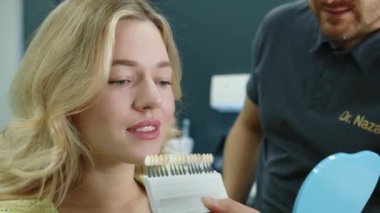 Diş gösteren, takma dişlerini seçen çekici genç bir kadın yakından çekilirken erkek dişçi muayenehanesinde diş implantı örnekleri sunuyor. Yüksek kalite 4k görüntü