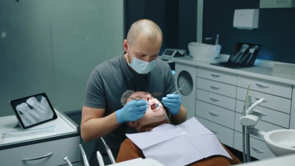 头戴医疗面罩的秃头牙医检查男性病人 用牙刷和舒适的牙科诊所的凹镜在半坐姿张大嘴 高质量的4K镜头 — 图库视频影像