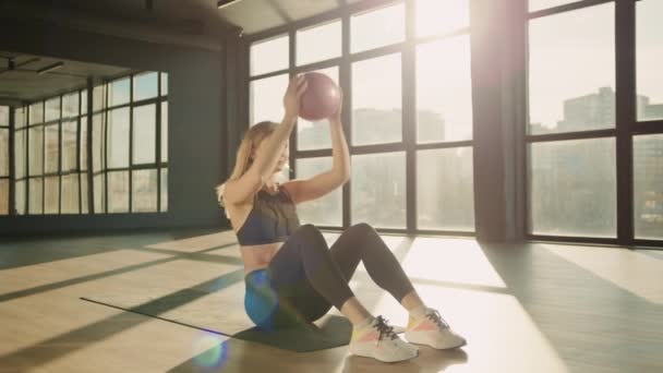 一个女孩手里拿着一个球 做仰卧起坐 在一个有巨大窗户的宽敞体育馆里锻炼 那里阳光灿烂 高质量的4K镜头 — 图库视频影像