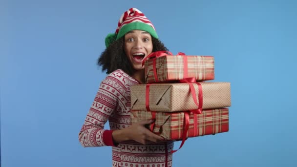 青い背景に立っているチェッカーと黄金の紙に包まれた3つのミドルサイズのボックスを保持するクリスマスの装飾とジャケットの若い幸せな美しさ 高品質の4K映像 — ストック動画