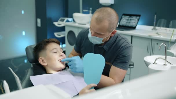 歯の状態を調べている医療マスクの歯科医が歯医者を診察している間 口を開いた開いた手の鏡で歯科医の椅子に座っている小さな男性患者は 問題を説明します 高品質の4K映像 — ストック動画
