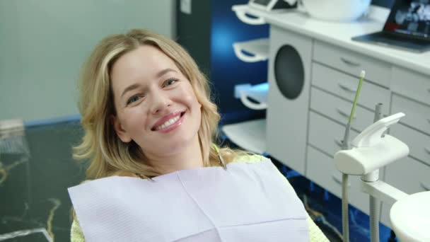 ホワイト家具を完備した居心地の良い歯科オフィスの胸に患者ナプキンが付いている半分座った位置のかなり陽気な女性の中型ショット 高品質の4K映像 — ストック動画