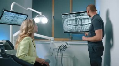 Dişlerinin röntgeniyle ekranın yanında duran ve dişçi koltuğunda oturan güzel sarışın kadına danışmanlık yapan, ışık saçmayan genç bir doktor. Yüksek kalite 4k görüntü