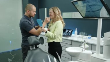 Genç erkek doktor ve güzel hasta ofiste, takma dişleri seçiyorlar, iletişim kuruyorlar, gülüyorlar. Beyaz mobilyalı rahat bir dolap, büyük ekran. Klinikte. Yüksek kalite 4k görüntü