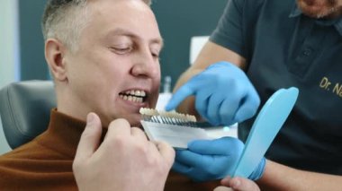 Tanımlanamayan erkek doktor, erkek hastaya diş implantı numuneleri sunuyor. Bir numuneyi koltukta oturan adamın dişlerine yerleştiriyor. Yüksek kalite 4k görüntü