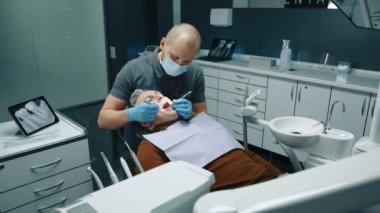 Tıbbi maskeli ortodontist, orta yaşlı erkek hastaları muayene eden bir tişört. Geniş ofiste dişçi aynası ve diş fırçasıyla modern ekipman kullanıyorlar. Yüksek kalite 4k görüntü