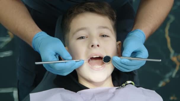 男性手戴乳胶手套 用牙刷 凹镜检查儿童下牙 病人平静地坐在牙医椅子上 胸前有餐巾 高质量的4K镜头 — 图库视频影像