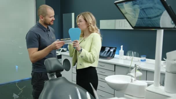 歯科インプラントのサンプルを提供する若い歯科専門家は 手の鏡 医師と患者が笑うかわいいブロンドの女性に 完全に装備された歯科オフィス 高品質の4K映像 — ストック動画
