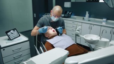 Diş matkabı ve ayna kullanan kel dişçi hastanın ağız boşluğunu temizliyor ve göğsünde peçeteyle oturuyor. Yüksek kalite 4k görüntü