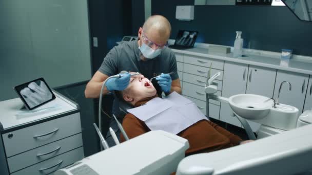 戴医用手套的秃顶专业牙医 使用牙钻和镜子对胸部餐巾患者口腔进行口腔卫生清洁 高质量的4K镜头 — 图库视频影像
