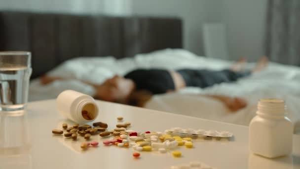 無意識の少女が部屋のベッドに横たわっている 前景では 多くの異なる錠剤が混沌と散らばっており 彼女が過剰な錠剤を服用したことを示しています 過剰摂取シナリオ 4Kについて — ストック動画