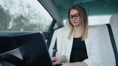 Bir iş kadını bir arabanın arka koltuğunda dizüstü bilgisayarda çalışır. Beyaz bir takım elbise ve gözlük takıyor, yüksek yoğunlukta ve dikkatle dizüstü bilgisayarların klavyesine yazıyor. Yüksek kalite 4k görüntü.