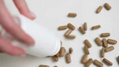 Kahverengi bitkisel hapların beyaz bir masaya dökülüşünü yakından çek. Bir el tarafından tutulan beyaz konteynırdan, doğal ilaç çözümleri sergileniyor..