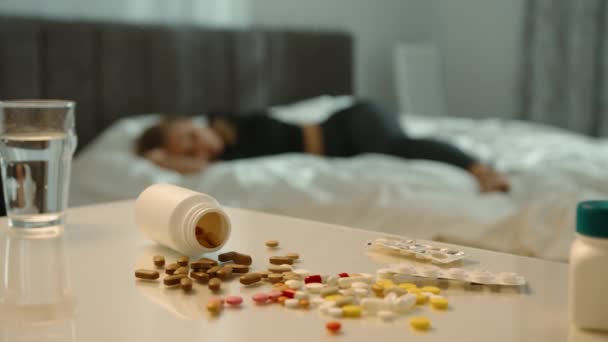 房间里的一幕 前厅有一张桌子 上面布满了各种药丸和瓶子 在背景中 一个女孩在床上昏迷不醒地躺着 突出了一个危急的情况 — 图库视频影像