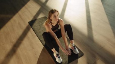 Sarışının biri antreman yapıyor, karın egzersizi yapıyor ve kollarını arkaya atıyor. Güneşli bir odada siyah bir paspasın üzerinde yatıyor. Spor ve sağlıklı bir yaşam tarzı. Üst Manzara. Yüksek kalite 4k görüntü.