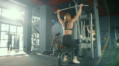 Geniş tutuş, dikey çekme egzersizi. Açık renk saçlı bir kız spor salonunda çalışıyor güneş ışığıyla yıkanmış büyük camlardan içeri akıyor. Yüksek kalite 4k görüntü.