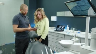 Genç atletik dişçi elinde diş implantı örnekleri tutuyor, ofiste gri duvarları ve beyaz mobilyaları olan güzel sarışına danışmanlık yapıyor. Yüksek kalite 4k görüntü