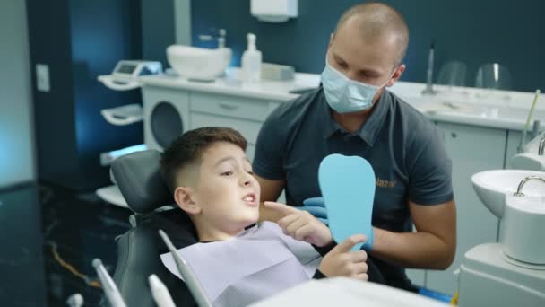 若い男性歯科専門家は 胸にナプキンと座って歯科医の椅子に座っている若い患者に 病気の歯を示す手の鏡を保持することを再確認します 高品質の4K映像 — ストック動画