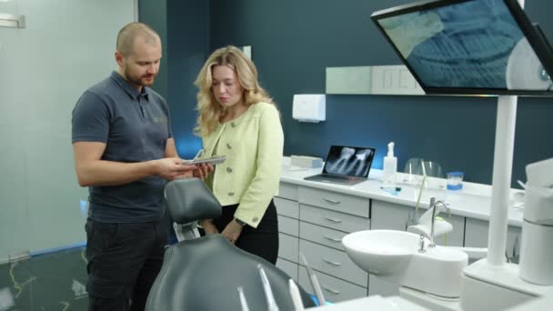 年轻的运动牙医拿着假牙样本 为站在办公室里的金发美女提供咨询 他们的办公室墙壁是灰色的 家具是白色的 高质量的4K镜头 — 图库视频影像