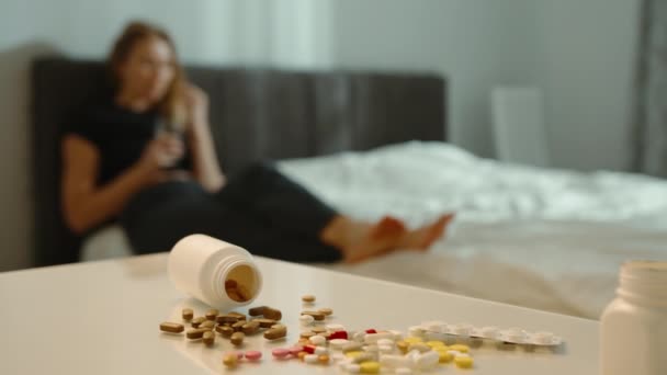 さまざまな錠剤やボトルが散らばった前景のテーブル付きのベッドルームシーン ぼやけた背景では 少女がベッドの上に座り 薬を飲んで うつ病状態を描きました — ストック動画