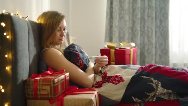 一个金发女郎躺在床上 身边都是圣诞礼物 她检查体温 发现她病了 咳嗽不止 假日病痛和康复的悲惨景象 — 图库视频影像
