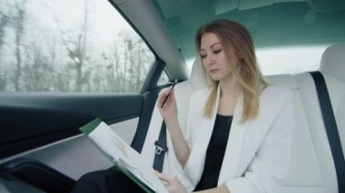 Beyaz ceketli açık renk saçlı bir kız arka koltuklarda iş çizelgeleriyle çalışıyor. O, elinde bir kalem tutuyor. Araba hareket ediyor. Pencerenin dışındaki hava bulutlu. 4k görüntü.