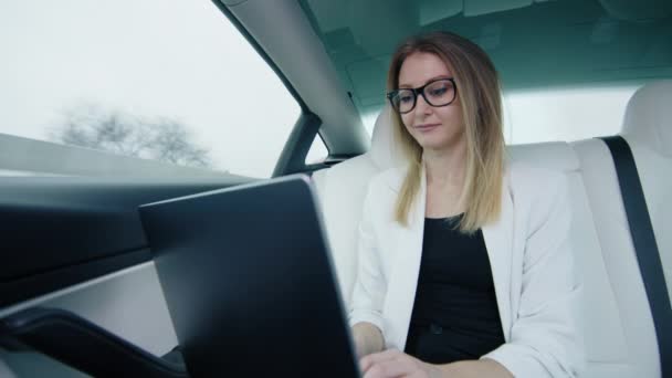 一个戴着眼镜和白色夹克的漂亮姑娘坐在汽车后备箱里专心致志地坐在笔记本电脑上 全神贯注地投入城市旅行中的工作 — 图库视频影像