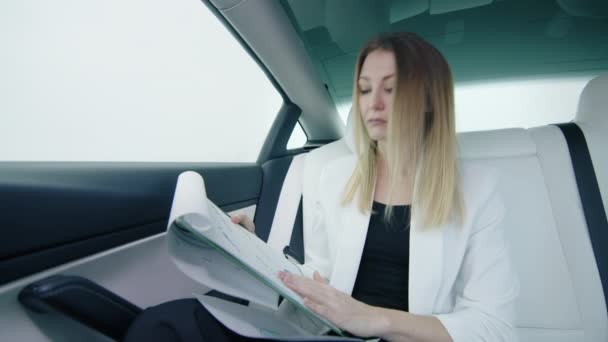 年次報告書を調べる電気自動車のビジネスウーマンは 注意深くチャートを見て 白いスーツを着て白い座席を持つ豪華な車に座りました 4K映像について — ストック動画