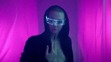 Sert görünüşlü bir kız, siber punk tarzı giyinmiş bir şekilde sanat alanında dans ediyor. Aydınlatma ve holograma sahip tekno gözlük takıyor ve siyah bir pelerin giyiyor..