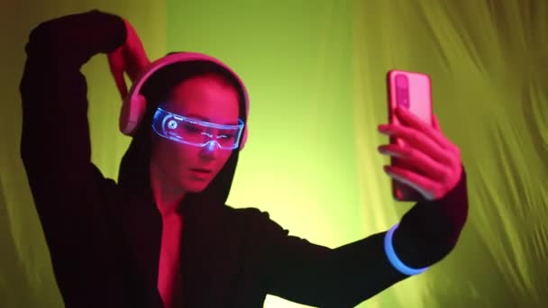 一个带着全息图和耳机的网络朋克女孩在一个五彩缤纷的空间里跳舞 随着光线的变化 她带着手机自拍 — 图库视频影像