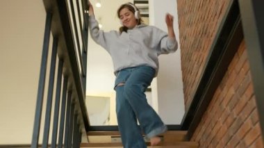 Kulaklıkla müzik dinleyen neşeli bir kız, apartmanının merdivenlerinden birinci kata kadar dans ediyor. İki katlı, çatı katı tarzı bir daire. Yüksek kalite 4k görüntü